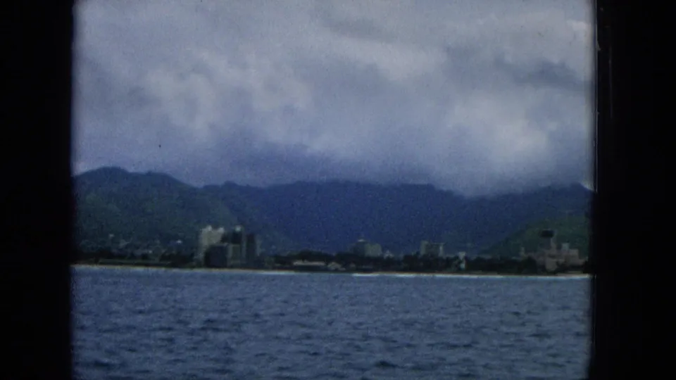 1965 Hawaii travel-film frame showing blue water, a distant shoreline with scattered buildings, green mountain slopes, and low clouds across the ridge.