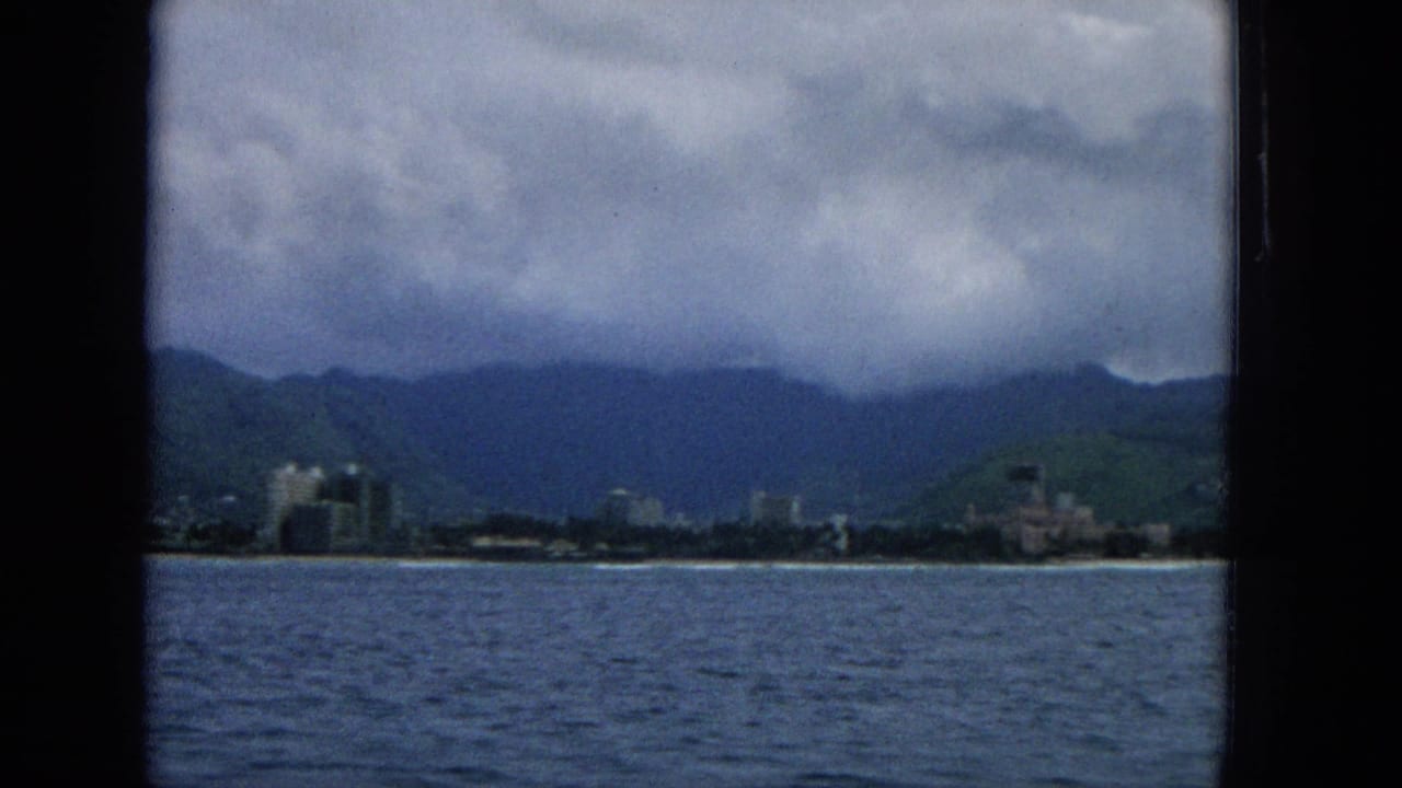 1965 Hawaii home-movie frame looking from across the water toward shoreline buildings, green mountains, and a heavy overcast sky.