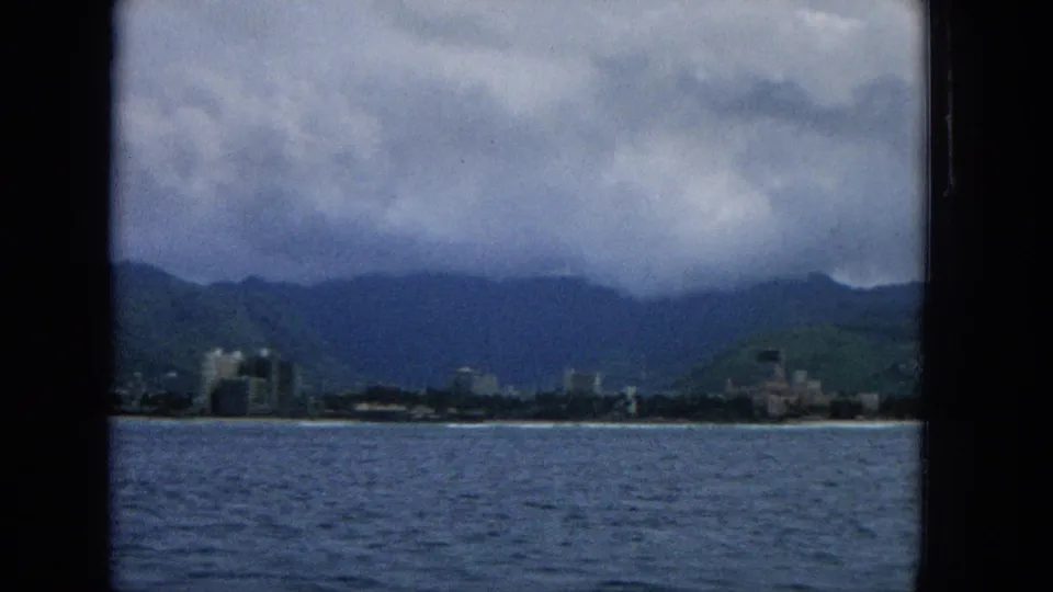 1965 Hawaii home-movie frame looking from across the water toward shoreline buildings, green mountains, and a heavy overcast sky.