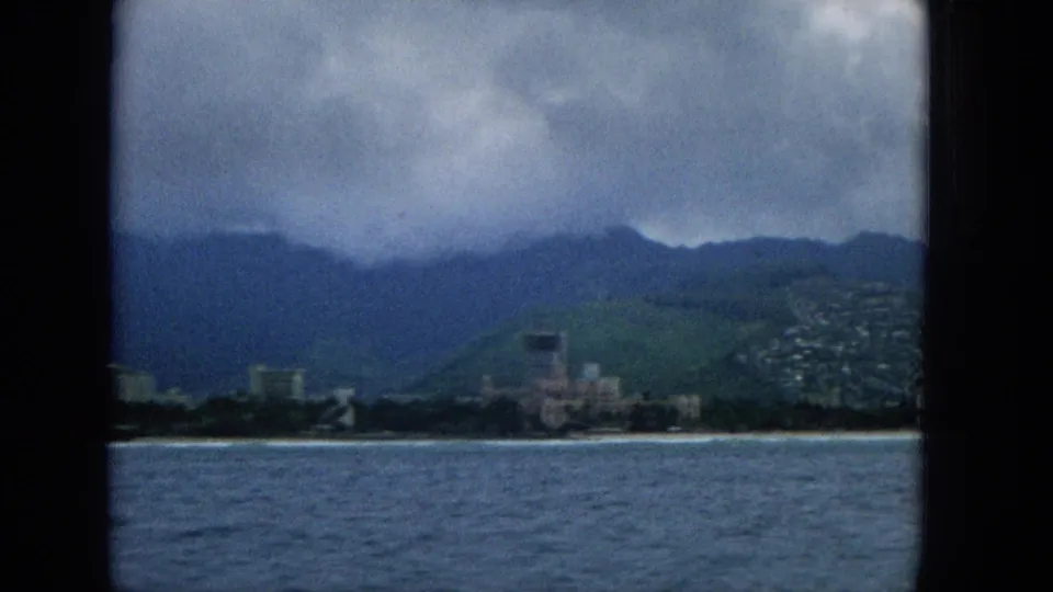 Offshore 8mm view of a Hawaiian coastal area with mid-century buildings, hillside homes, green slopes, and low clouds.