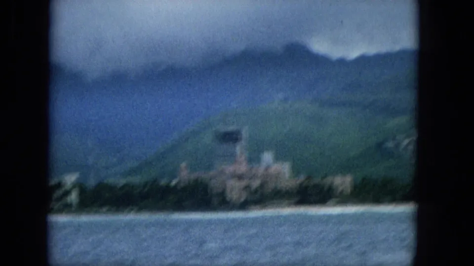 Soft 8mm frame from 1965 Hawaii showing ocean in the foreground, a developed shoreline, green hillside, and mist-covered mountains.