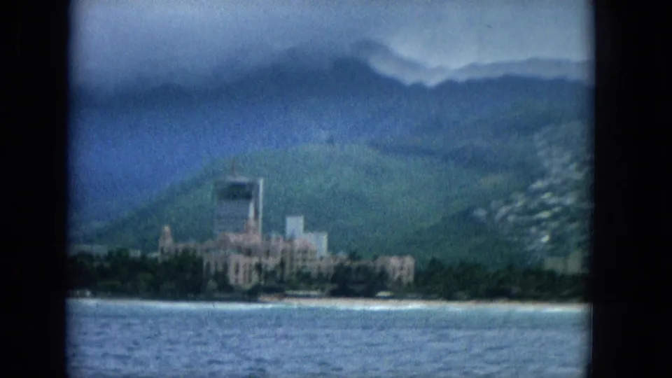 1965 Hawaii travel footage frame showing a coastal building cluster beneath green mountains, filmed from a boat across blue water.