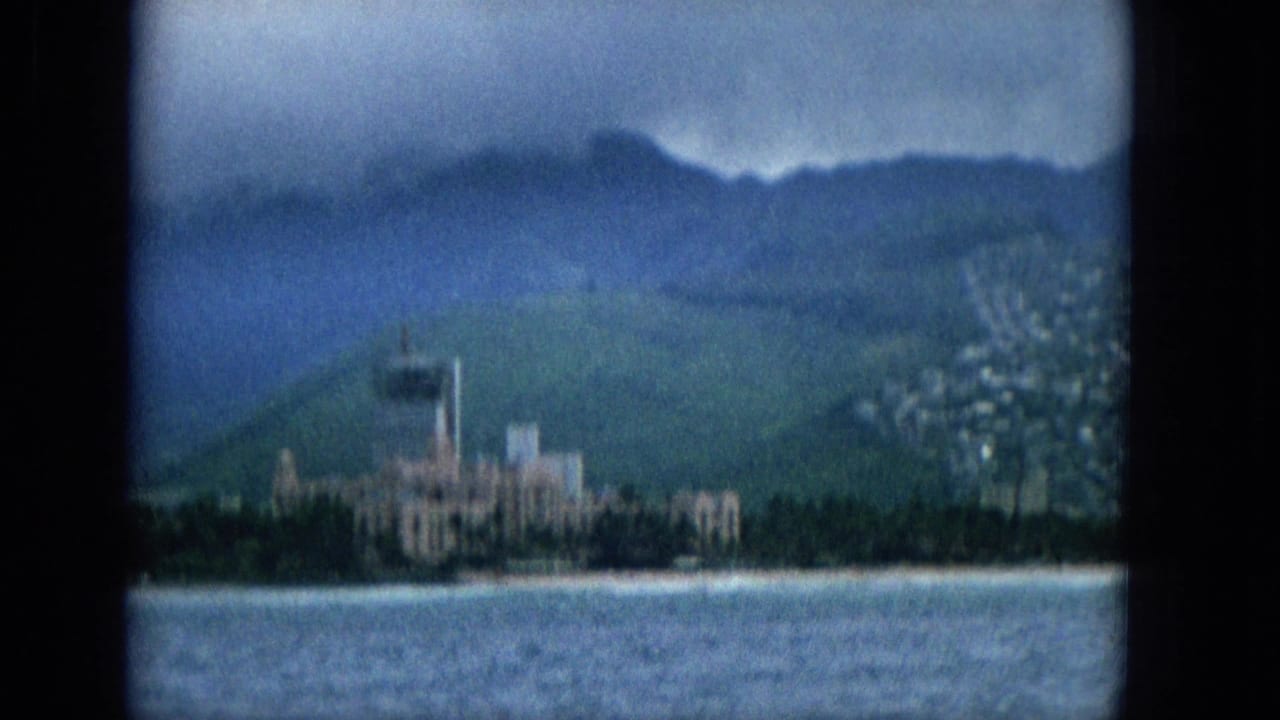 1965 home-movie frame from offshore showing a Hawaiian coastal strip with ocean in the foreground, beachside buildings, a prominent distant tower, green hills, and soft overcast light.