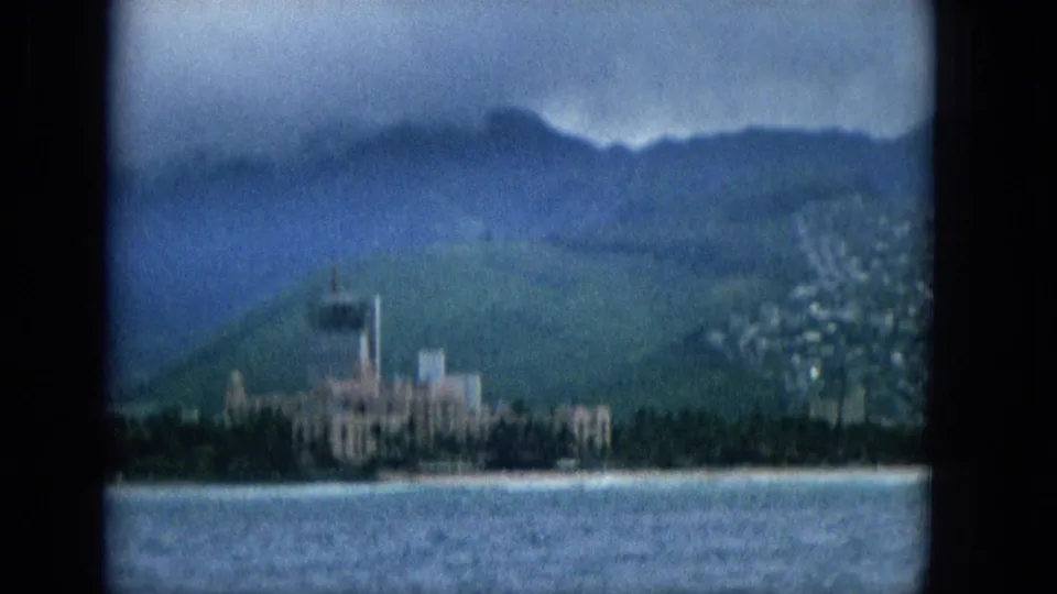 1965 home-movie frame from offshore showing a Hawaiian coastal strip with ocean in the foreground, beachside buildings, a prominent distant tower, green hills, and soft overcast light.