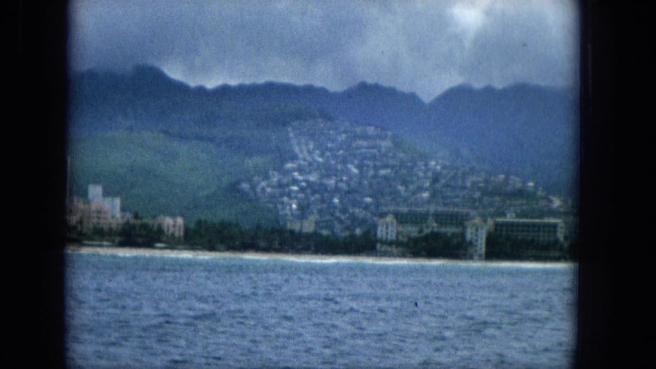 1965 offshore view of a Hawaiian coast with resort-like buildings along the beach, hillside housing behind them, calm water in front, and a cloudy mountain backdrop.
