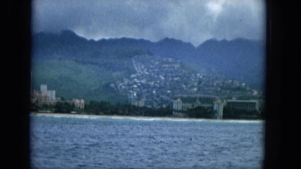 1965 offshore view of a Hawaiian coast with resort-like buildings along the beach, hillside housing behind them, calm water in front, and a cloudy mountain backdrop.