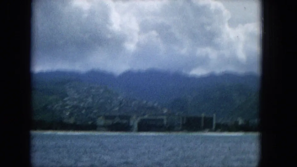 1965 Super 8 frame showing a Hawaiian coastal panorama from a boat, with rippled water, a narrow beach, low shoreline structures, green hills, and dense clouds above.