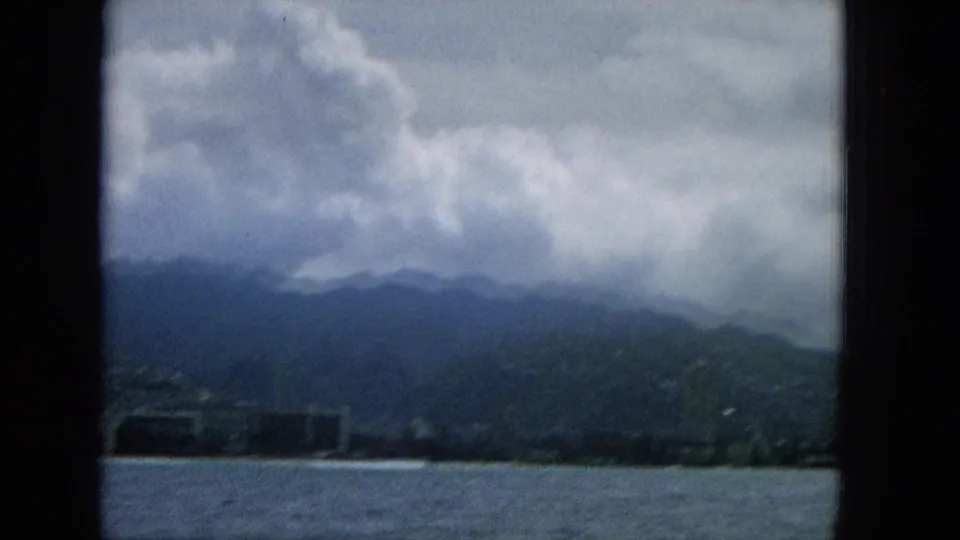1965 home-movie view from offshore looking toward a darker Hawaiian hillside or mountain ridge, with ocean in front, low coastal structures, and thick overcast clouds.