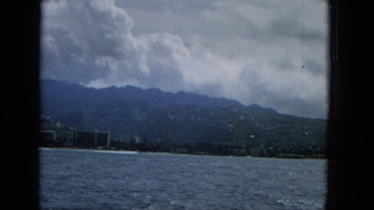 1965 Hawaii coastal city view with choppy water, a distant shoreline, mountain ridge, and storm clouds framed by dark window bars.