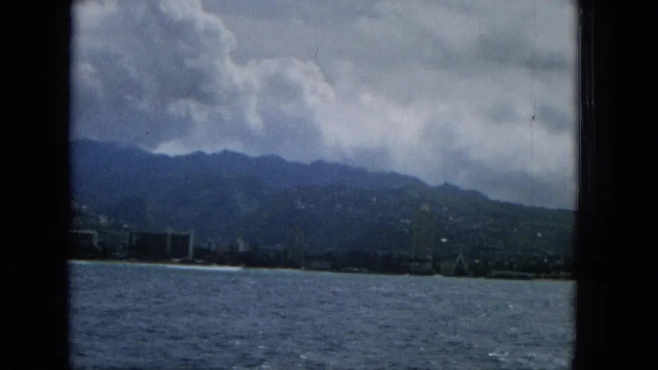 1965 Hawaii coastal city view with choppy water, a distant shoreline, mountain ridge, and storm clouds framed by dark window bars.