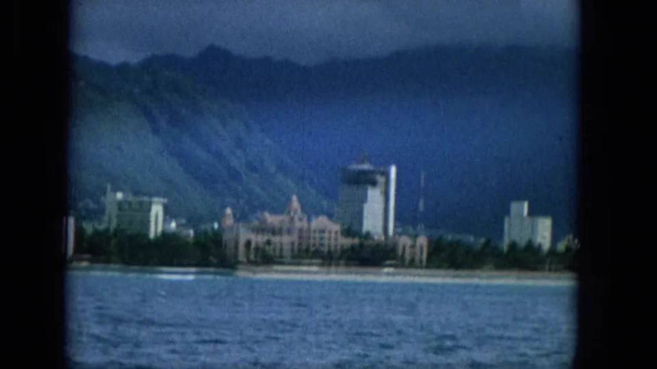A closer 1965 Hawaii shoreline view shows beachfront buildings, a taller tower, blue water, and green mountains under darkening sky.