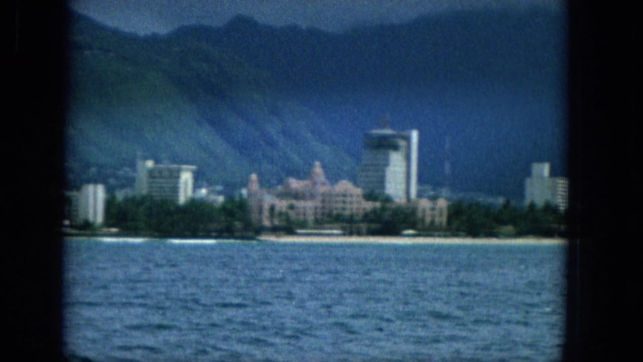 1965 Super 8 frame showing an offshore view toward the Oahu shoreline, with ocean water, a pale beach, waterfront buildings, and dark mountains under clouds.