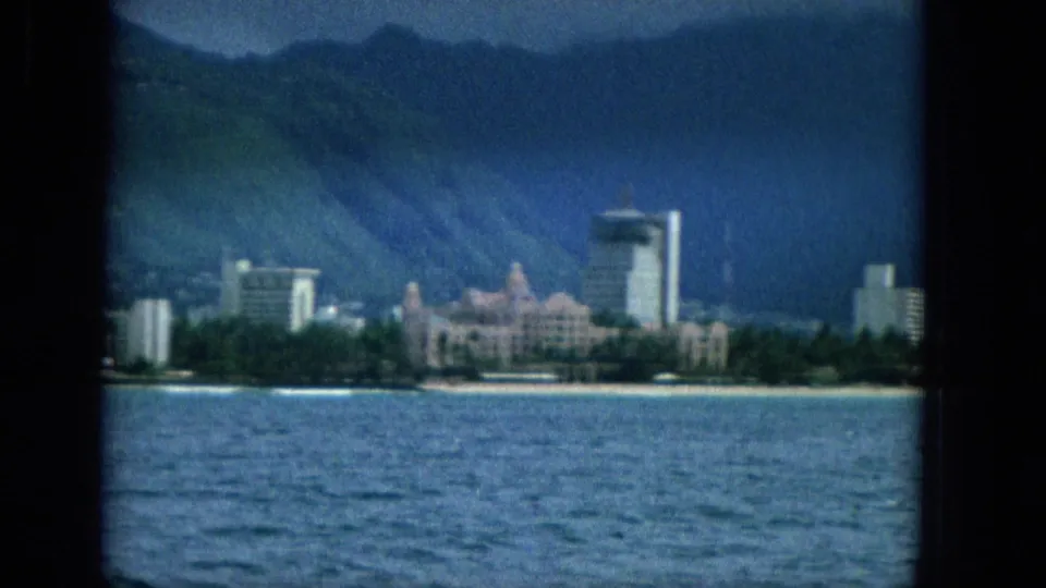 1965 Super 8 frame showing an offshore view toward the Oahu shoreline, with ocean water, a pale beach, waterfront buildings, and dark mountains under clouds.