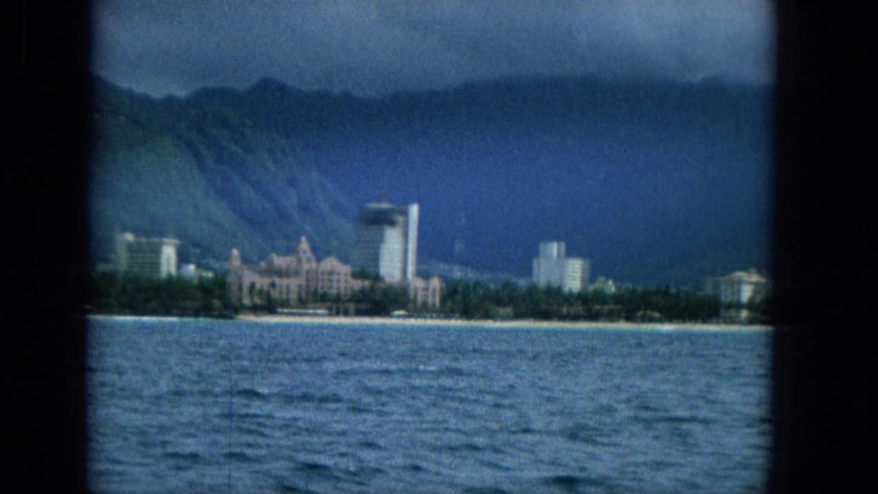 Super 8 frame from the 1965 Hawaii shoreline view, showing ocean in the foreground, clustered waterfront buildings, a tall tower, and green mountains in the background.