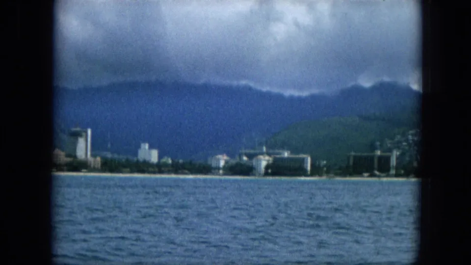 1965 Super 8 frame showing a darker section of the Oahu shoreline, with ocean water, low waterfront structures, taller buildings, and clouded mountains behind.