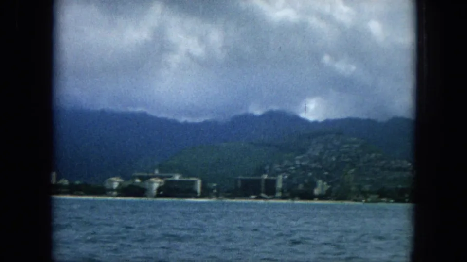 Super 8 frame showing ocean foreground and a developed shoreline below green hills and dark mountains, with heavy clouds above the ridge.