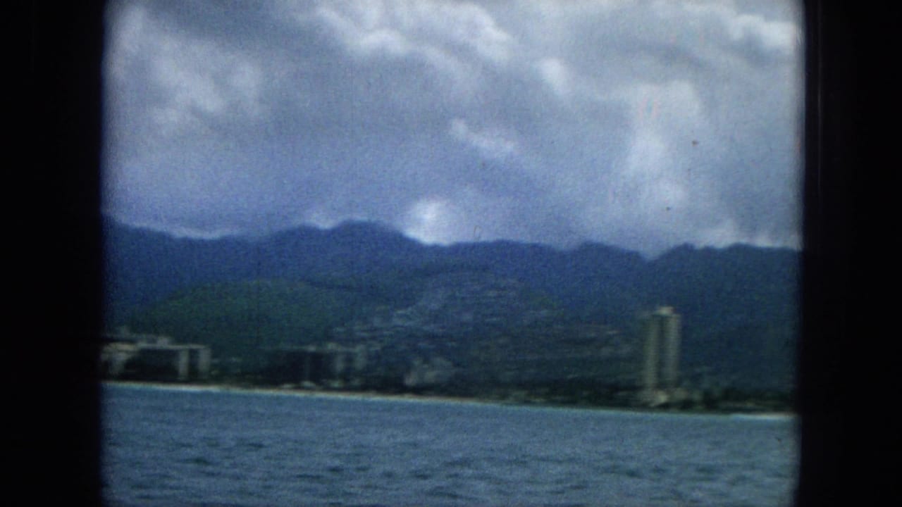 1965 home-movie frame showing a Hawaiian coastal city viewed from the water, with low shoreline buildings, a tall tower, mountain ridges, and dark clouds.