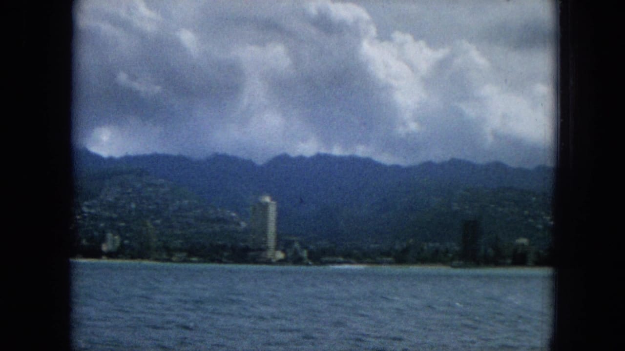 Waterfront skyline with a tall shoreline tower, ocean foreground, and mountains in the distance.