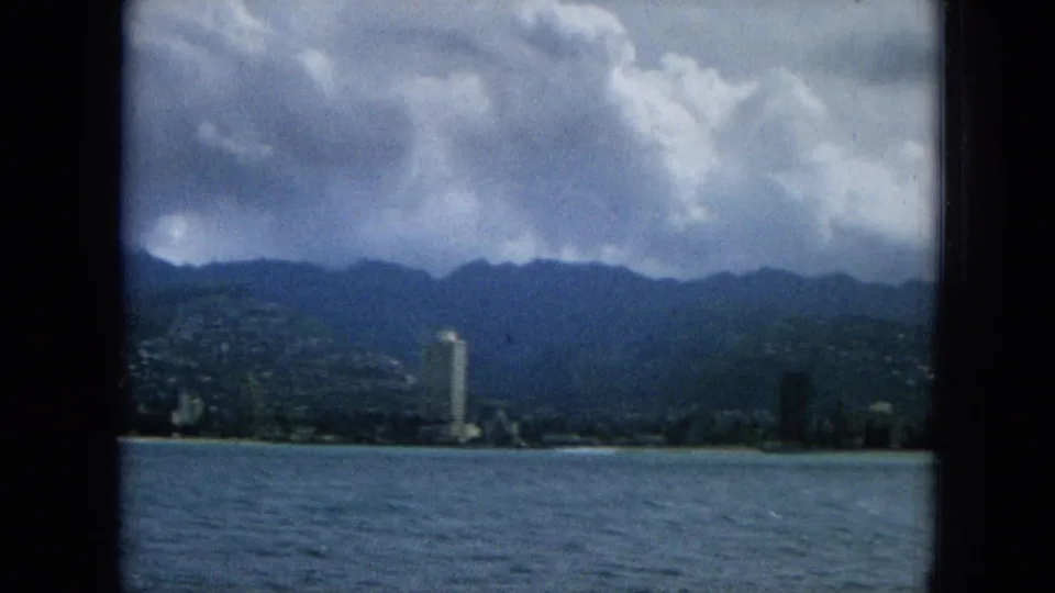 1965 home-movie view from a boat toward a Hawaiian waterfront skyline, with a prominent high-rise tower, distant hillside development, mountains, and heavy clouds.