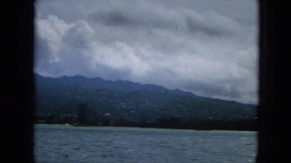 1965 home-movie frame of a Hawaiian shoreline seen from the ocean, with darker buildings near the coast, mountain slopes behind, and overcast sky above.