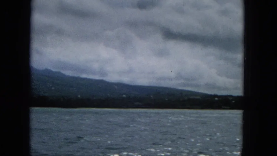 1965 home-movie frame looking from a boat across water toward a low Hawaiian shoreline and broad mountain slope under a dense cloudy sky.