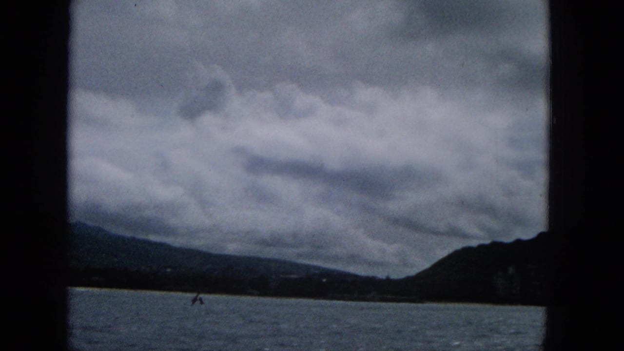 1960s 8mm frame looking from a boat across water toward a dark mountain ridge and cloudy coastal horizon, with faint shoreline structures visible.