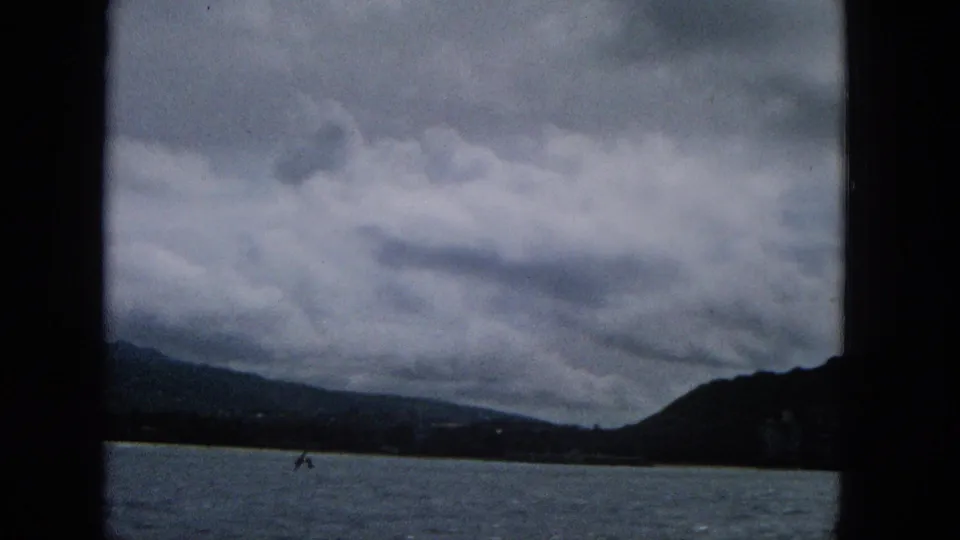 1960s 8mm frame looking from a boat across water toward a dark mountain ridge and cloudy coastal horizon, with faint shoreline structures visible.