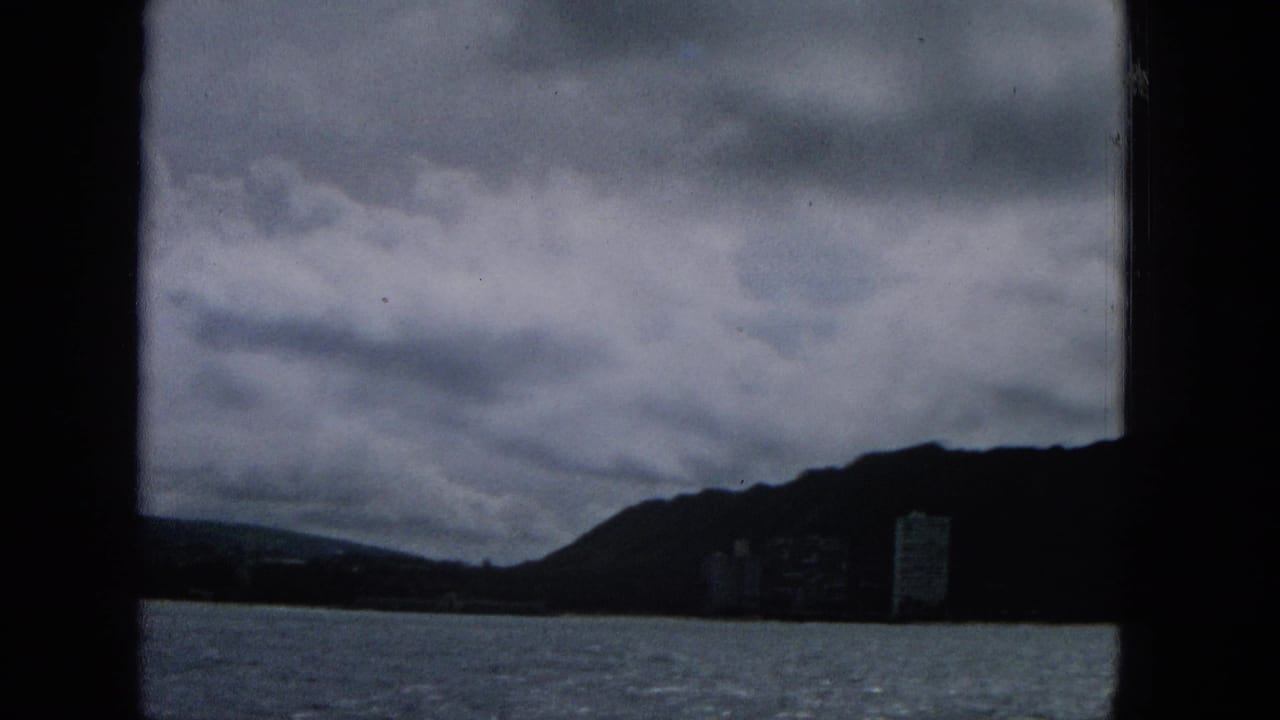 Cloudy coastal water view with a mountain slope and distant tower-like building.