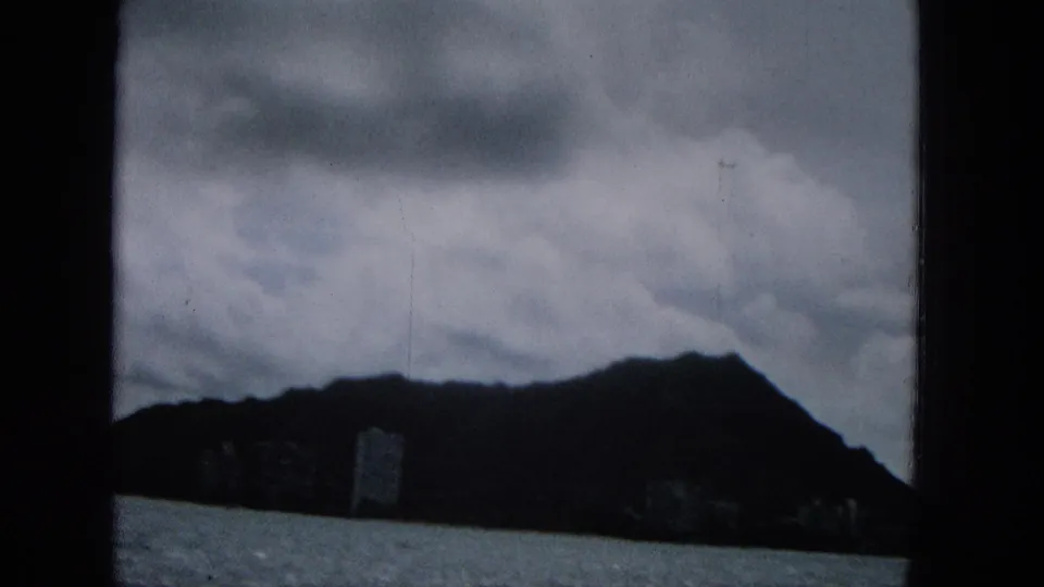 1960s 8mm frame with soft focus and dark exposure showing water, a mountainous coastal edge, low clouds, and distant buildings near the shore.