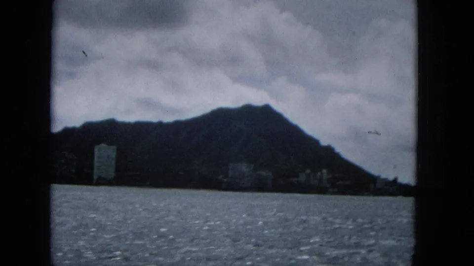 1960s 8mm frame from the boat view showing rippled water, a dark mountain profile, low clouds, and a cluster of distant coastal buildings including a tower-like form.