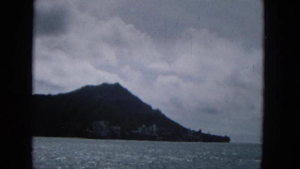 1965 8mm frame showing a dark coastal hill, distant shoreline structures, choppy water, and a cloudy sky, with film-frame borders visible.