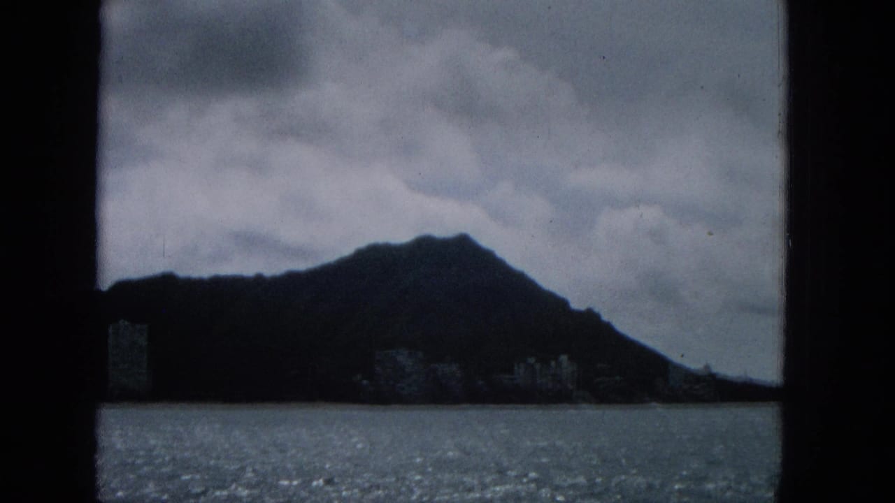 1965 8mm frame with a dark hill silhouette above the ocean, low clouds overhead, and faint shoreline buildings near the waterline.