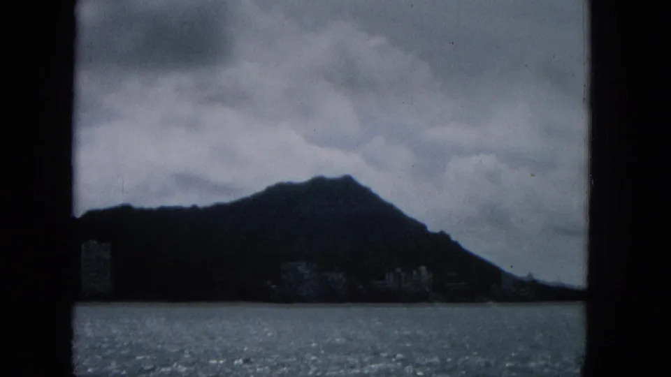 1965 8mm frame with a dark hill silhouette above the ocean, low clouds overhead, and faint shoreline buildings near the waterline.