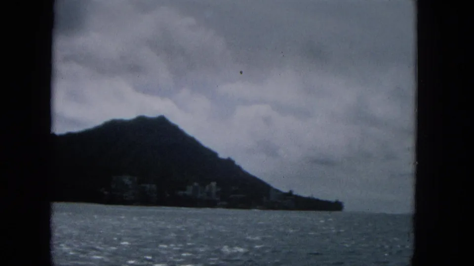 1965 8mm frame showing the side of a dark coastal hill, a small cluster of distant buildings, textured ocean water, and gray cloud cover.