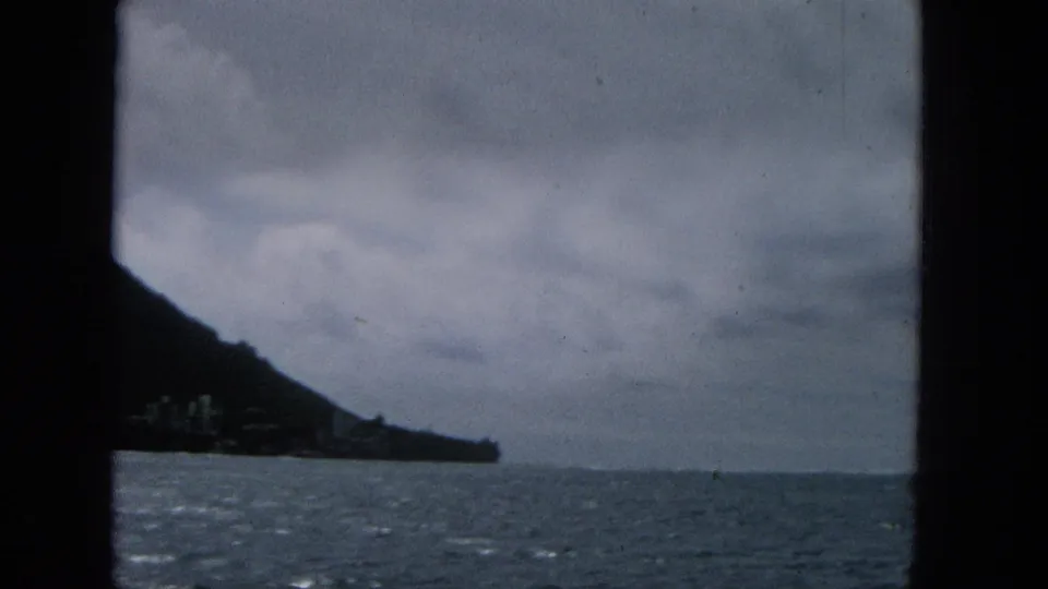 1965 8mm frame with choppy ocean in the foreground, a dark hillside at left, a low shoreline point, and heavy clouds across the sky.