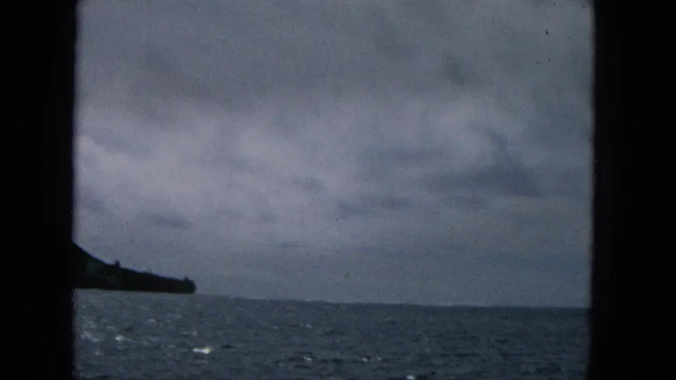 1965 Super 8 frame from a moving boat showing open ocean, low clouds, and a dark coastal point at the left edge, likely off Molokai based on clip metadata.
