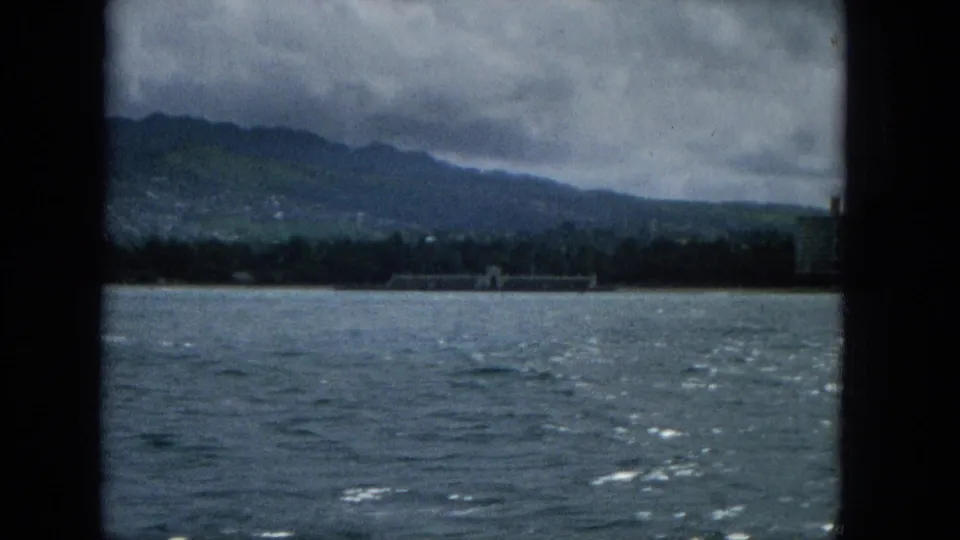 1965 Super 8 coastal boat view with rippled ocean, a distant shoreline, and mountain slopes under heavy cloud cover, likely Molokai according to metadata.