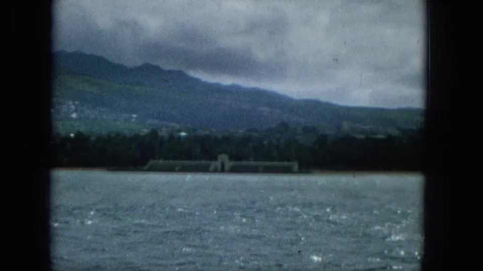 1965 Super 8 frame showing a muted coastal landscape from offshore, with water in front, trees along the shore, and hazy mountains under overcast sky.