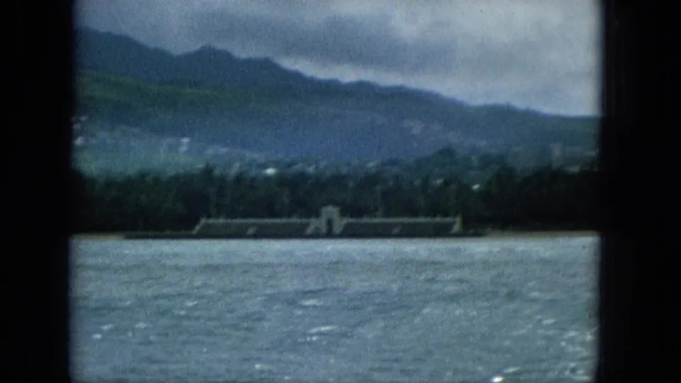 1965 Super 8 boat-view frame with choppy water, a distant low coastal structure or wall, tree cover, and dark island mountains beneath clouds.