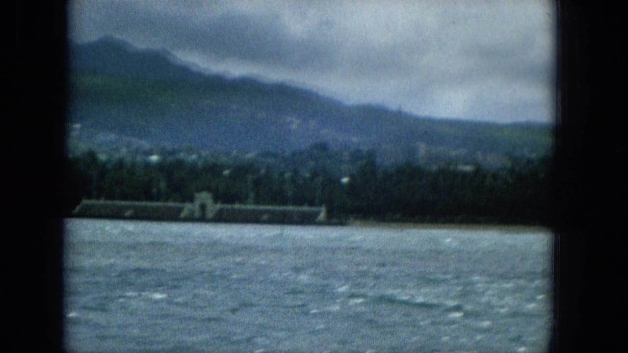 1965 Hawaiian coastal view from a boat, showing rippled water, a palm-lined shoreline, a long low waterfront structure, and hazy mountains beneath overcast sky.