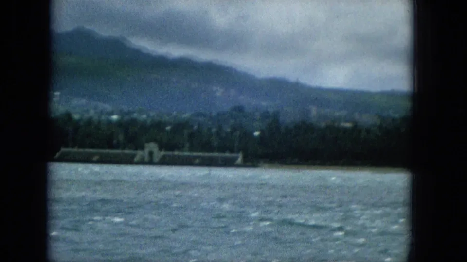 1965 Hawaiian coastal view from a boat, showing rippled water, a palm-lined shoreline, a long low waterfront structure, and hazy mountains beneath overcast sky.