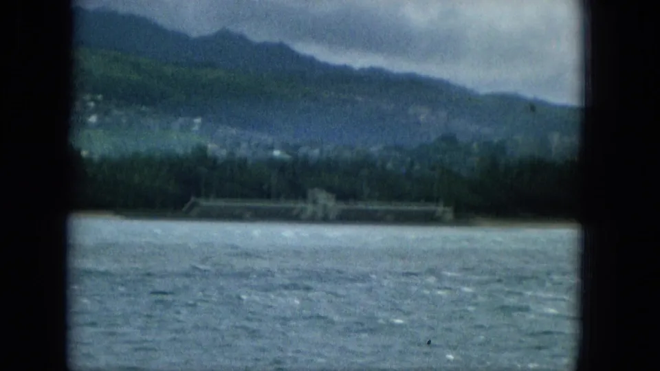 1965 home-movie frame looking from the water toward a Hawaiian shoreline with trees, a long low waterfront structure, and layered mountains in soft focus.