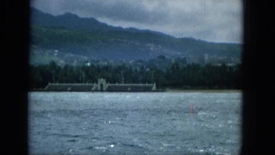 1965 Hawaiian shore filmed from a moving boat, with choppy water in the foreground, palms and a long waterfront building mid-frame, and blue-green mountains beyond.