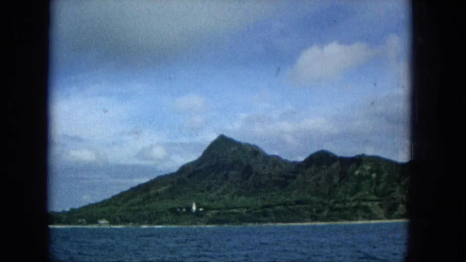 1965 Hawaiian island view across blue water, showing a steep green mountain peak, low shoreline, small light structure, and partly cloudy sky.