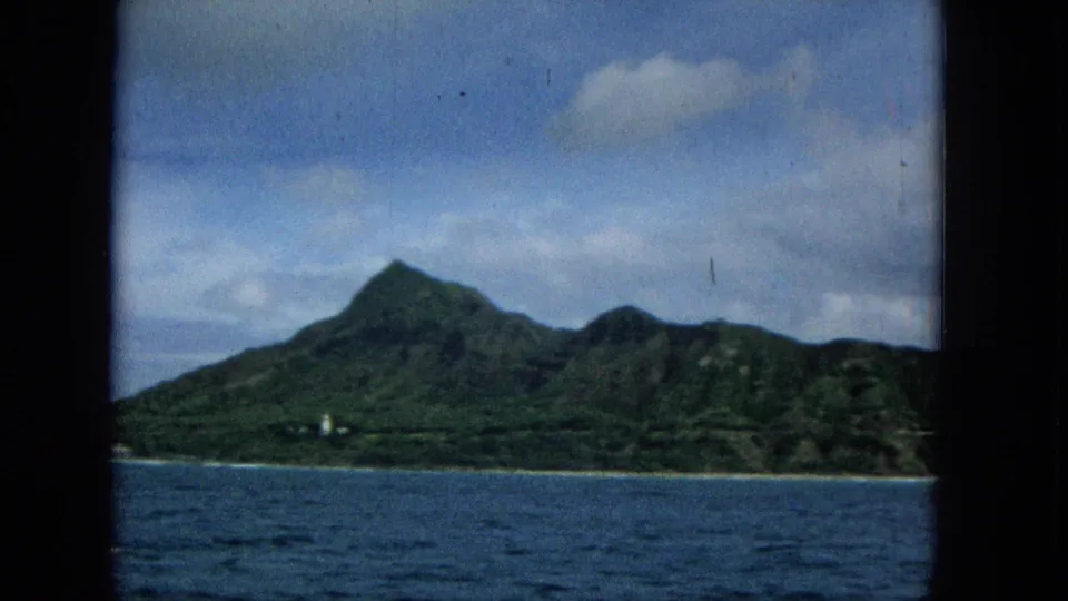 1960s Super 8 frame showing a green mountain shoreline across blue ocean, viewed through dark film-frame edges from an offshore vantage.