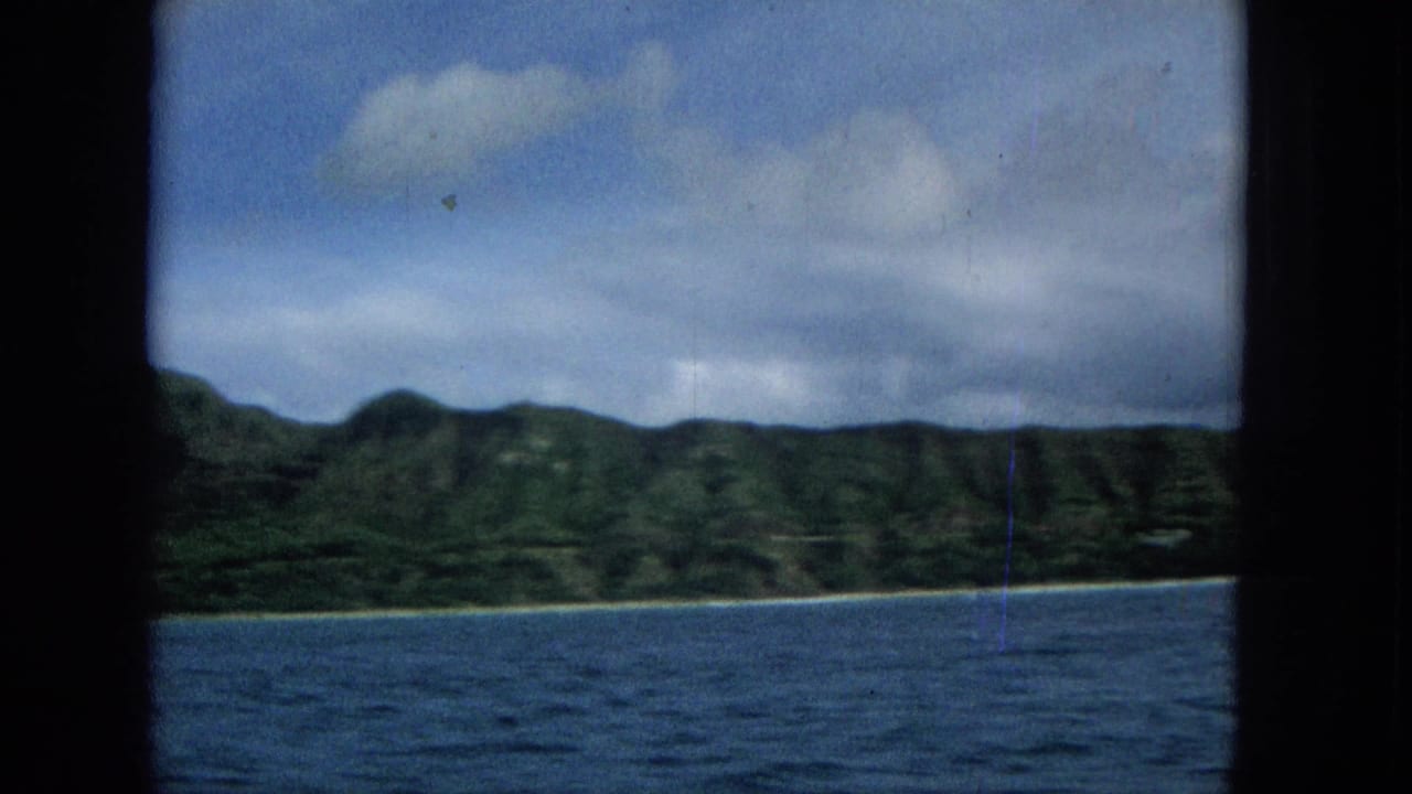 Offshore view of a green ridged coastline and narrow beach line, with blue water in the foreground and a dark vertical film or camera obstruction near center.