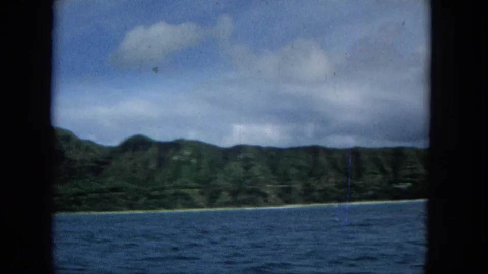 Offshore view of a green ridged coastline and narrow beach line, with blue water in the foreground and a dark vertical film or camera obstruction near center.
