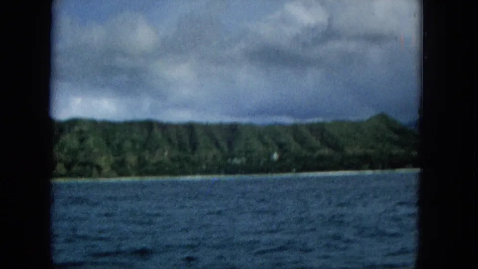 Super 8 frame of low green coastal hills beyond choppy blue water, with a pale shoreline and cloudy sky suggesting a moving boat perspective.