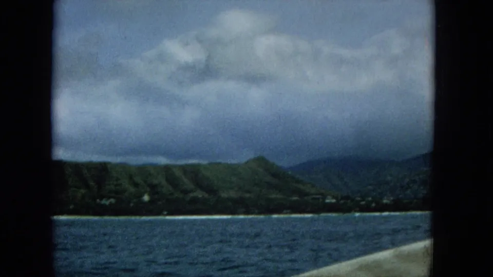 Offshore Super 8 view of a green mountain shoreline under layered clouds, with blue water in front and a light boat edge visible at the lower right.