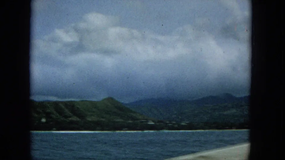 1965 8mm view toward Molokai coastline from a boat window, showing blue water, green hills, low shoreline, and heavy clouds.